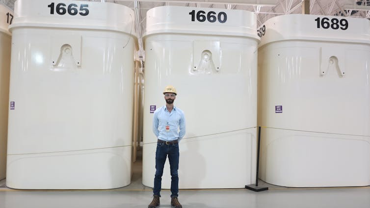 A man wearing a hard hat stands in front of large white cylindrical metal containers