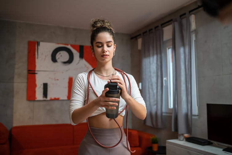 A young woman in gym clothes holds a supplement shaker bottle in her hands.