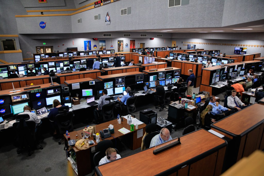 Dozens of engineers work at computer terminals in a wide shot of Firing Room 1.