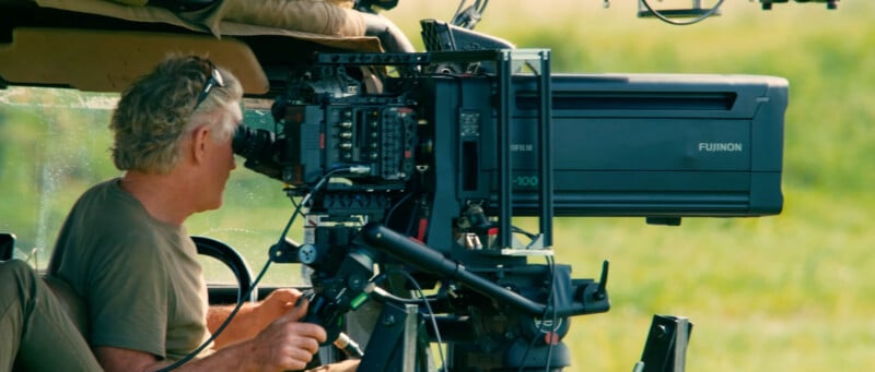 A person operates a large Fujinon broadcast camera, aiming it out of an open vehicle window, with green grass visible outside.
