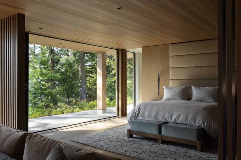 Modern bedroom with wood-paneled walls and ceiling, large glass doors open to a forest view, bed with white linens, and a neutral area rug on the floor.