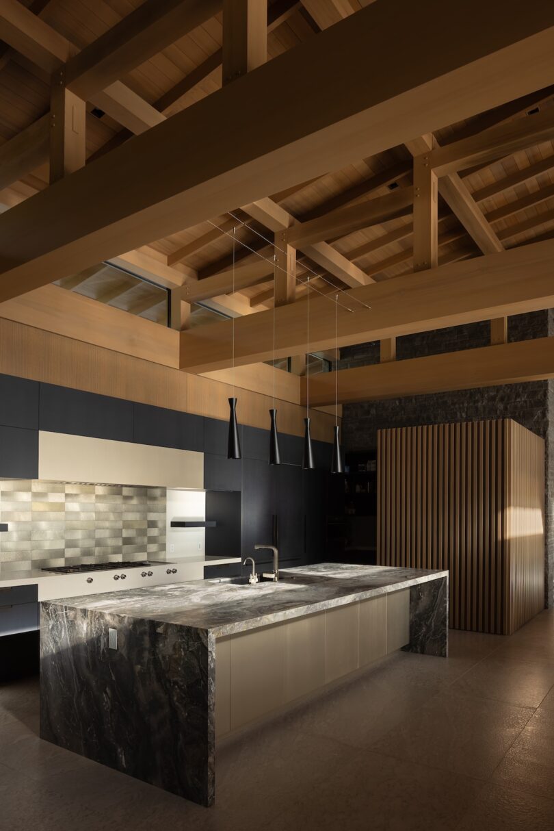 Modern kitchen with a large marble island, built-in appliances, dark cabinetry, tile backsplash, pendant lights, and exposed wooden ceiling beams.