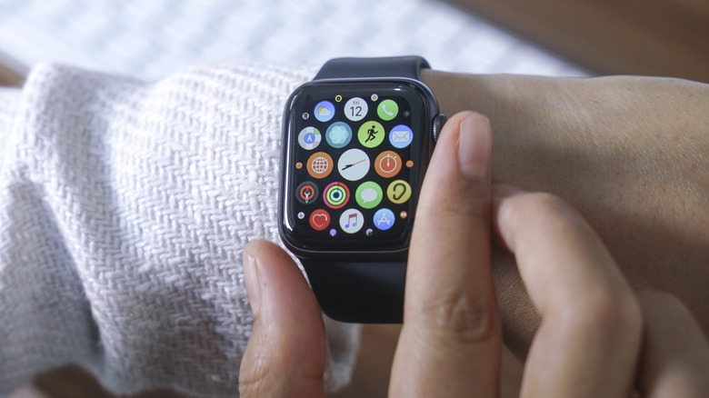 A closeup of an Apple Watch on an individual's left wrist, the thumb and forefinger of their right hand is around the watch as it displays a list of apps in a honeycomb pattern
