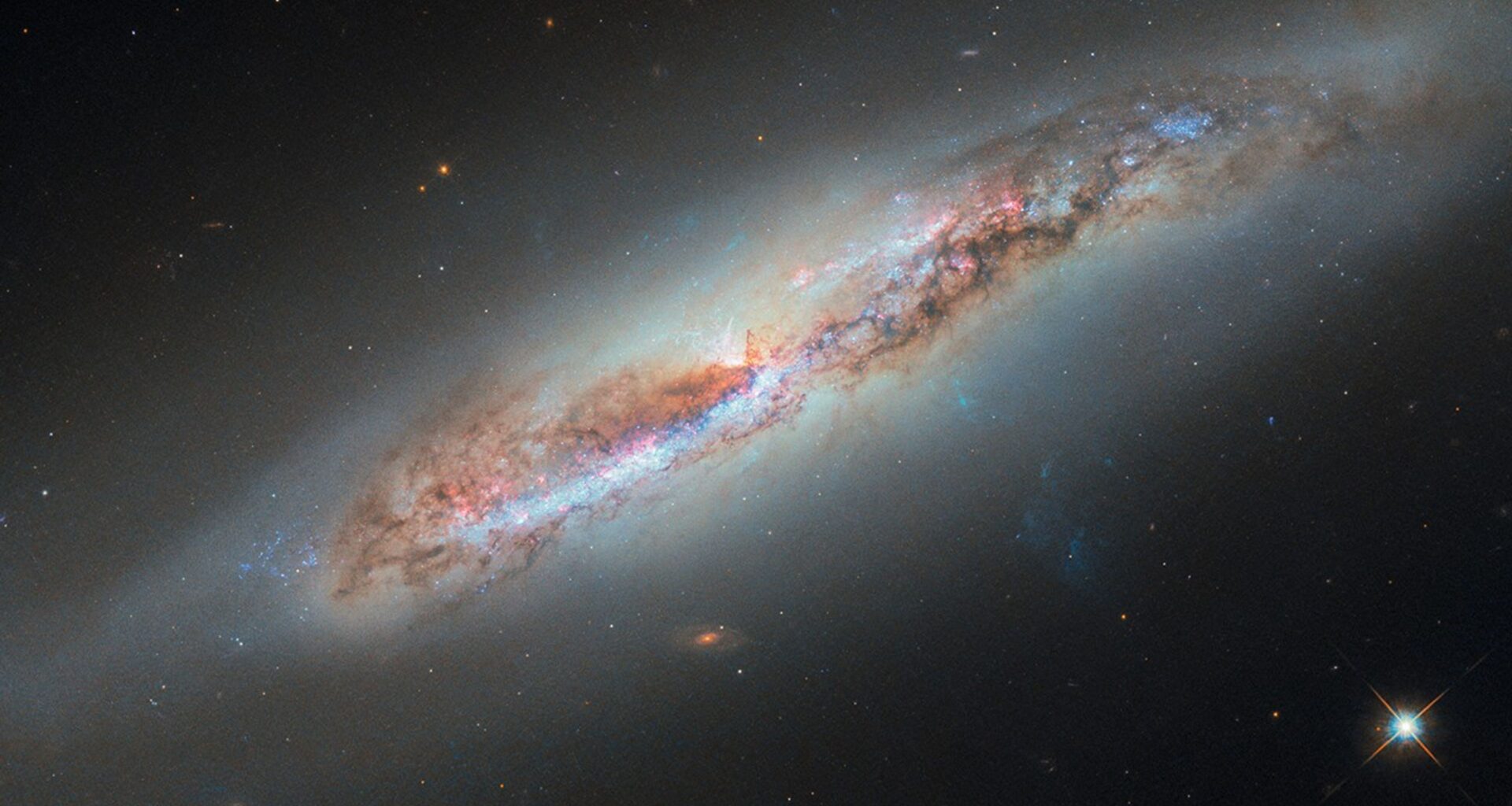 A nearly edge-on spiral galaxy. Its disk holds pink light from star-forming nebulae and blue light from clusters of hot stars. Thick dark clouds of dust block the strong white light from galaxy’s center. A faint, glowing halo of gas surrounds the disk, fading into the black background of space. A bluish plume of gas also extends from the galaxy’s core extending toward the lower-right corner of the image.