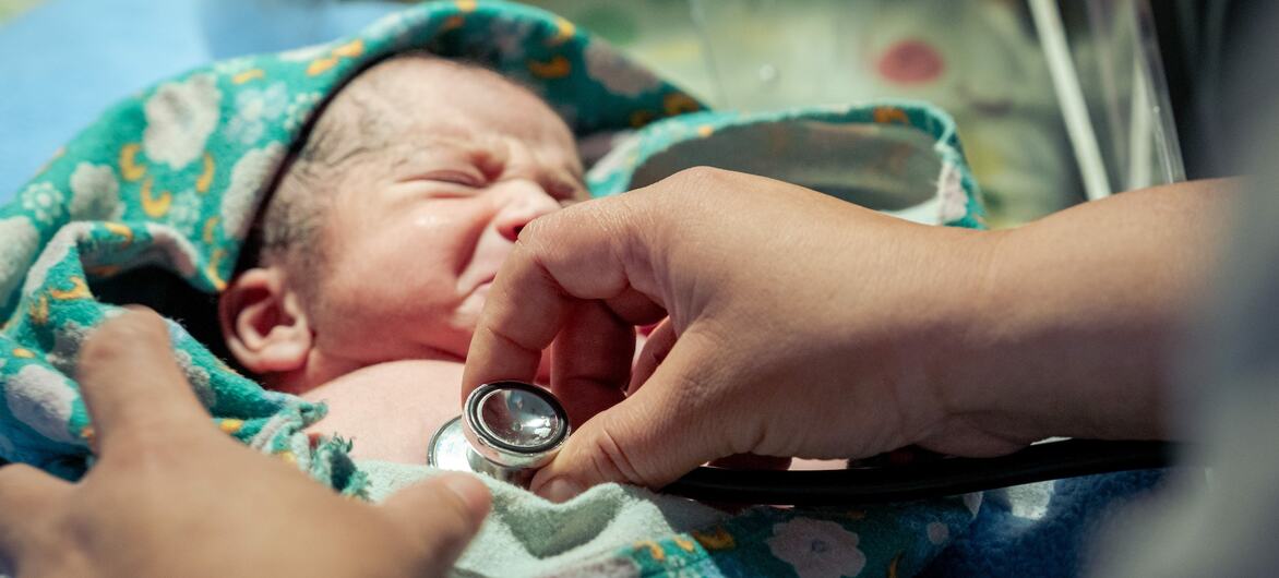 A close-up of a newborn baby wrapped in a patterned blanket, being examined with a stethoscope by a healthcare professional in a hospital setting.