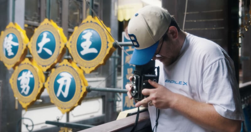 A man in a white shirt and baseball cap uses a vintage camera on a balcony, with ornate yellow and blue Chinese signs visible in the background.