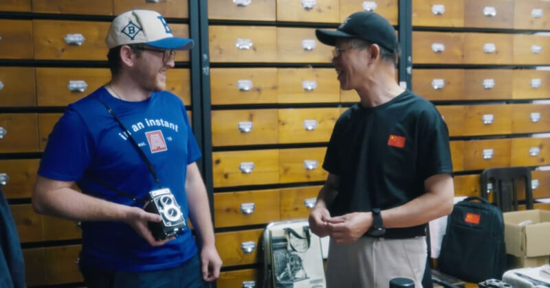 Two men stand and smile at each other in front of a wooden cabinet with many drawers. One man holds a vintage camera, and both wear casual clothes and caps. Various items are displayed on a table beside them.