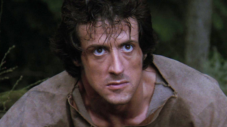 Sylvester Stallone's John J Rambo looks determined as he looks up while standing in a forest in First Blood