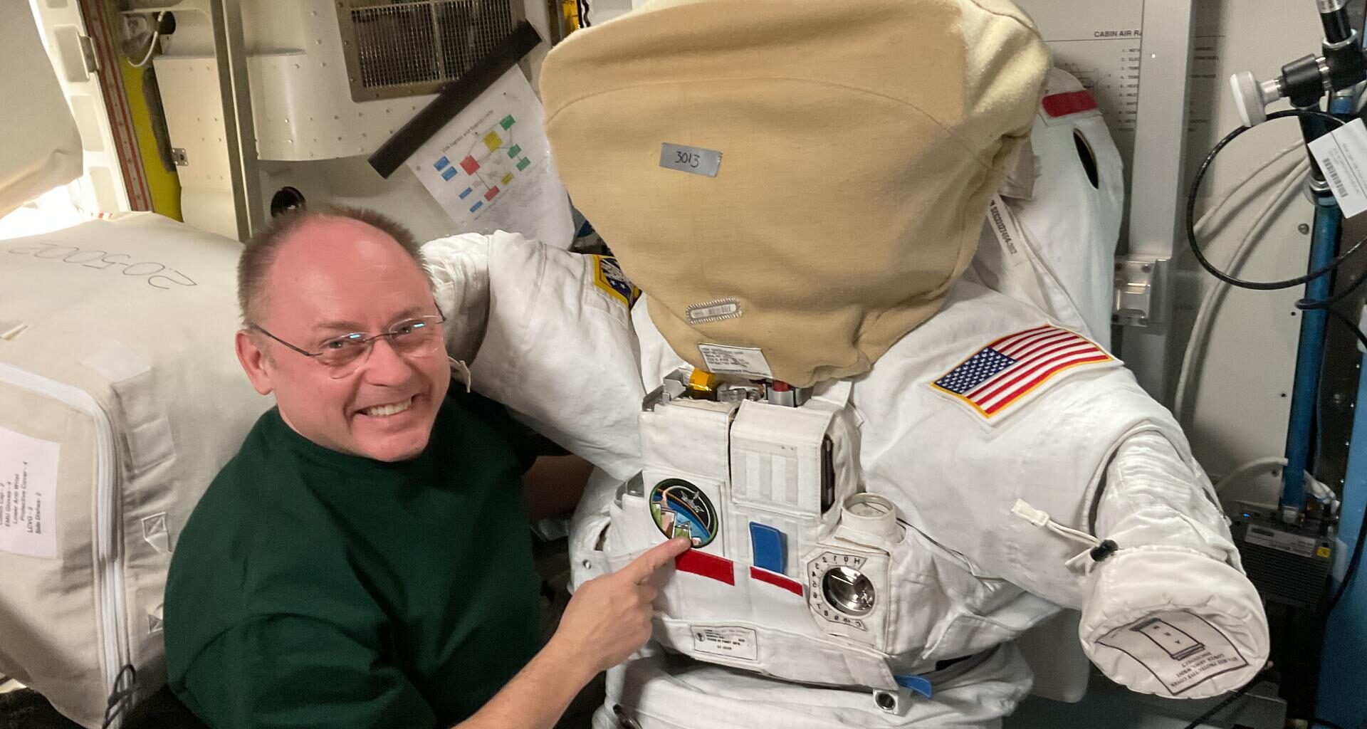 NASA astronaut and Expedition 74 Commander Mike Fincke poses inside the International Space Station’s Quest airlock next to a spacesuit. The helmet is secured with a protective cover designed to prevent scratches and contamination when the suit is not in use, ensuring the visor remains clear for spacewalks.