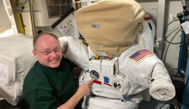 NASA astronaut and Expedition 74 Commander Mike Fincke poses inside the International Space Station’s Quest airlock next to a spacesuit. The helmet is secured with a protective cover designed to prevent scratches and contamination when the suit is not in use, ensuring the visor remains clear for spacewalks.
