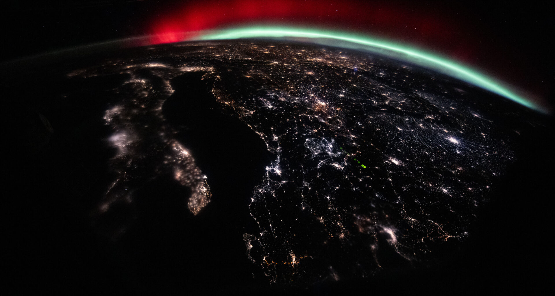 A green and red aurora streams across Earth’s horizon above the city lights of Europe.