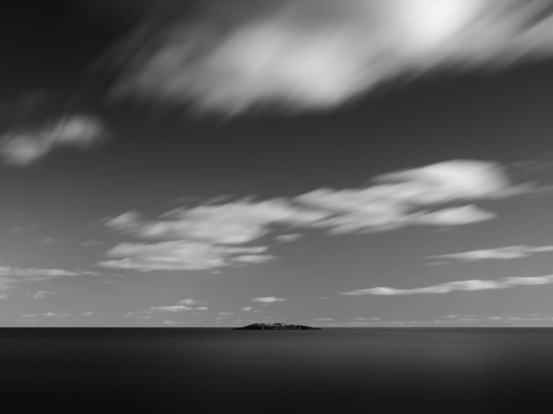 A small, rocky island sits alone on a calm sea beneath a sky streaked with fast-moving, blurred clouds in a black-and-white photograph. The scene appears peaceful and minimalistic.