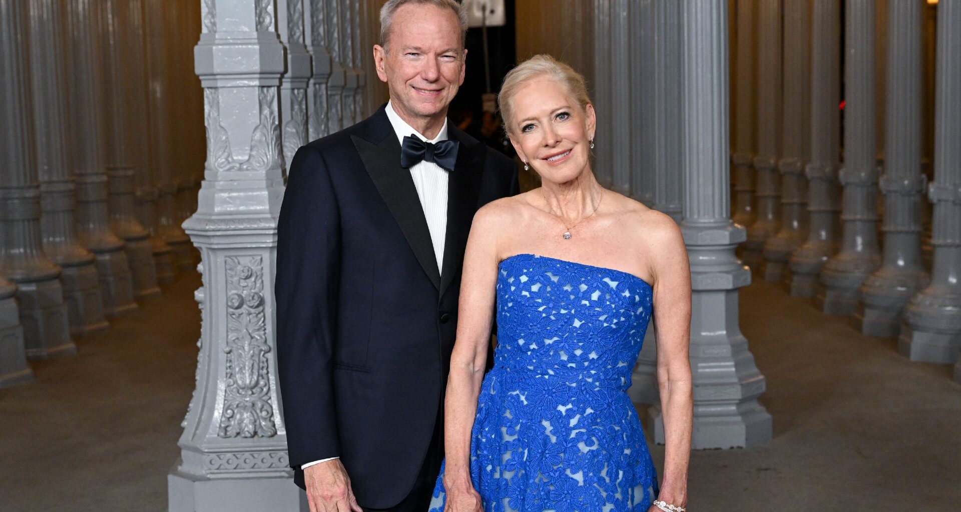 A man wearing a black suit and bowtie stands to the left of a woman in a sparkly blue dres.