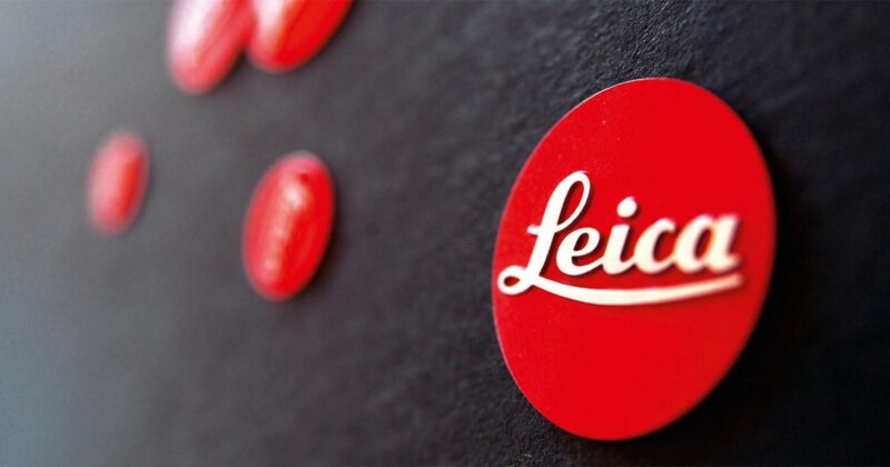 A close-up photo of multiple red circular badges on a black surface, with the frontmost badge displaying the white cursive "Leica" logo. The background badges are out of focus.