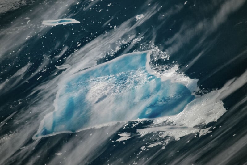Aerial view of a floating ice sheet surrounded by dark blue water and streaks of thin clouds, with small fragments of ice nearby.