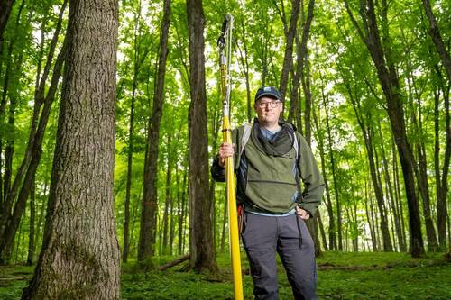 A man in a blue cap, glasses, and an olive-green jacket with a mesh neck cover holds a tall yellow pole. He stands in a vibrant green forest, looking forward with a slight smile.