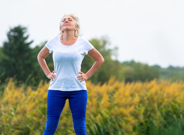 Adult woman with hands on hips looking upwards while wearing blue pants and white tee shirt with blurry yellow flowers in background