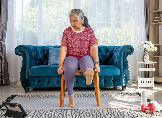 Senior woman sitting on a wooden chair, raising leg to stretch muscles and knees, Training exercise online with tablet In Living Room During Quarantine