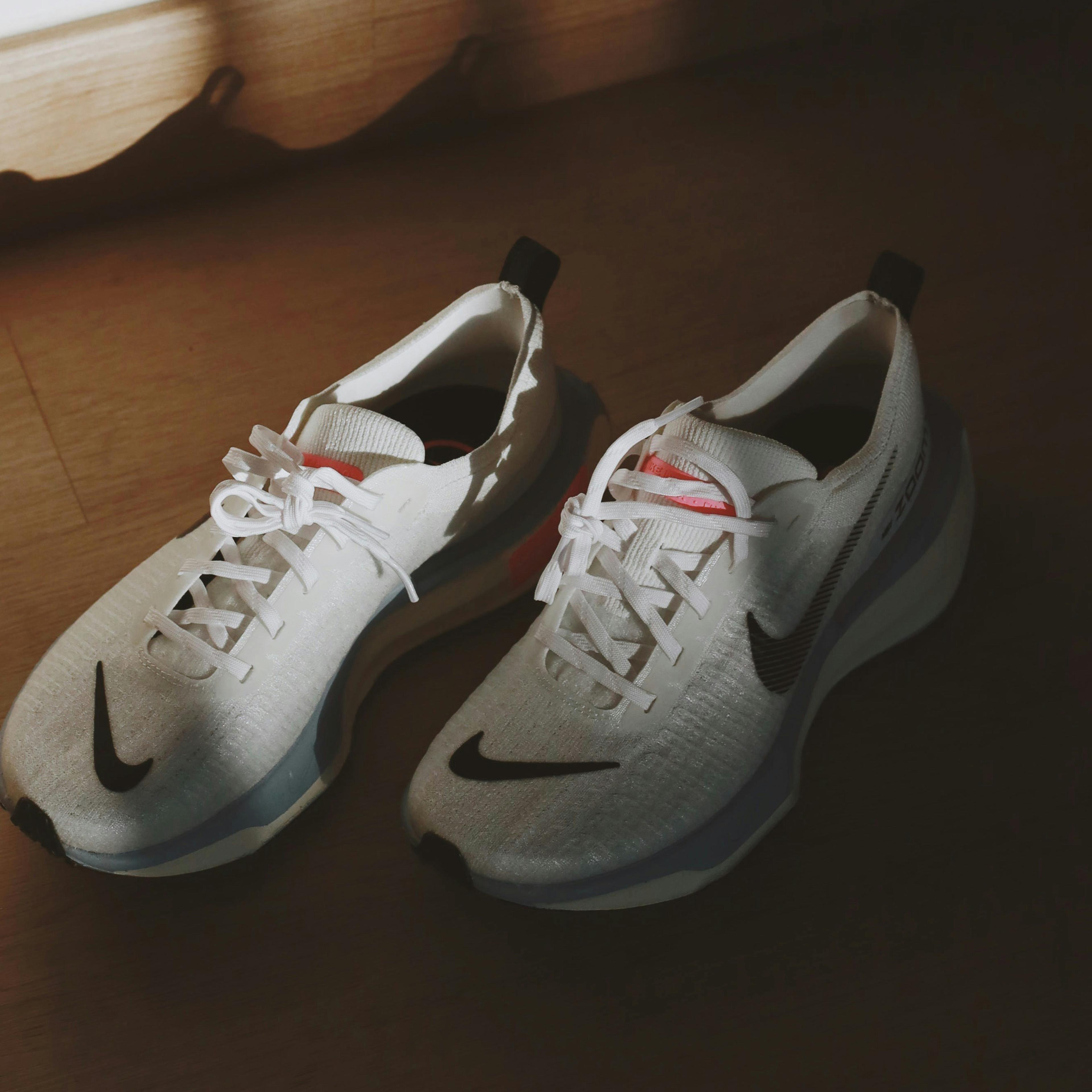 A pair of white trainers with a black tick Nike logo against a wooden floor, sunlight casting a shadow behind.