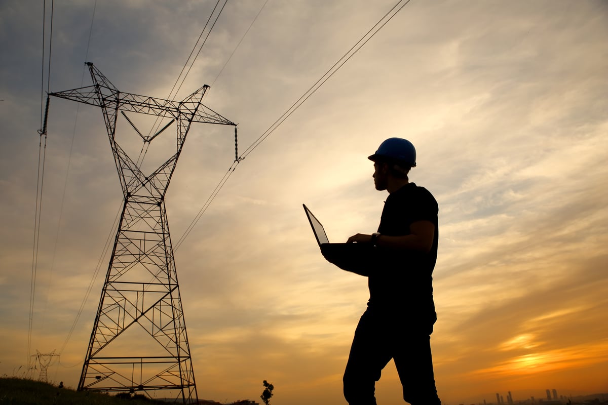 A person wearing a hardhat and holding a laptop near a power line.
