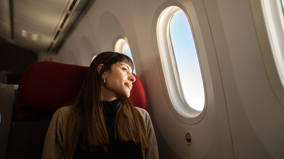 Woman seated by an airplane window, looking outside during a daytime flight.