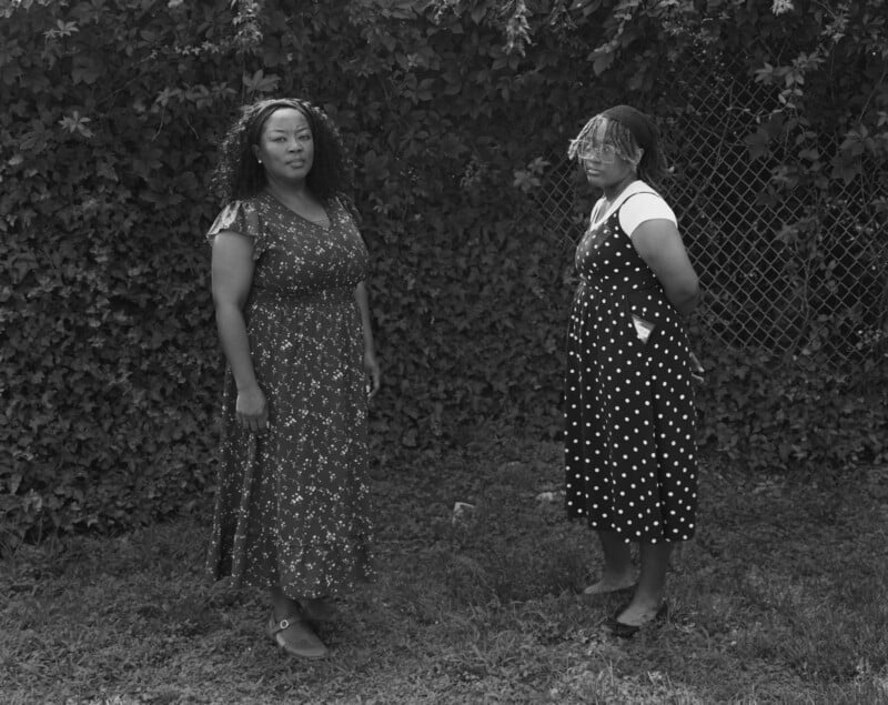 Two women stand on grass in front of a leafy, vine-covered fence. One wears a patterned dress and looks at the camera; the other, in a polka-dot dress and braids, stands with hands behind her back, looking slightly away.