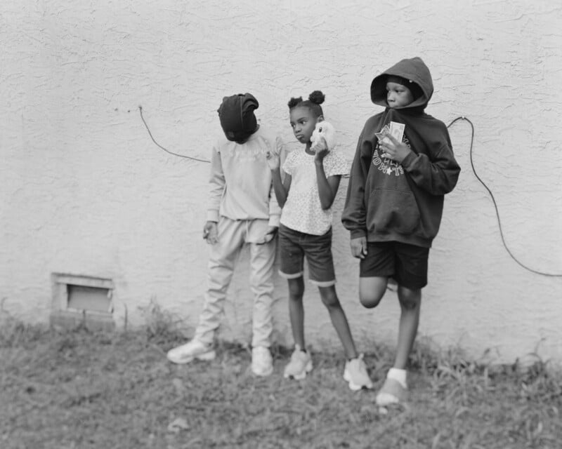 Three children stand against a textured wall. One child wears a hoodie and holds money, another has a shirt over their head, and the third, in shorts, holds a stuffed animal. Grass covers the ground in front of them.