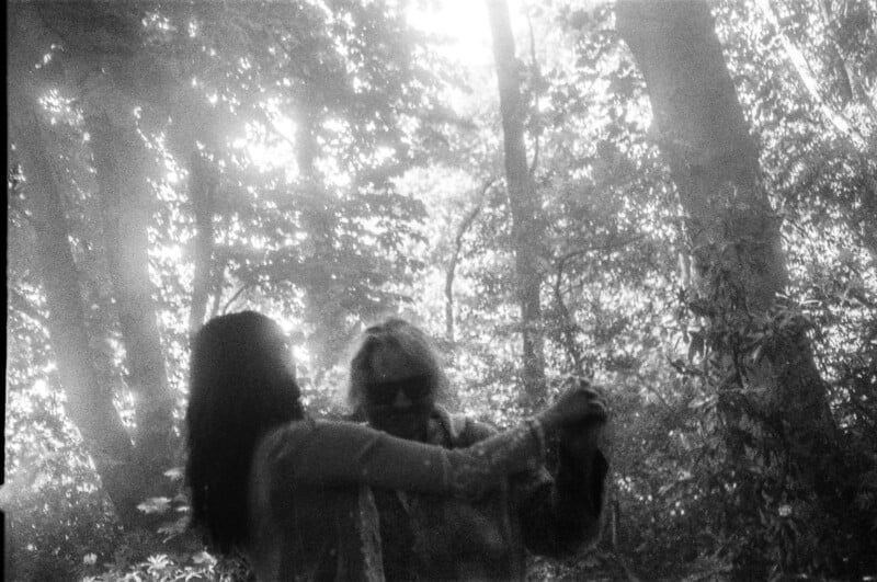 Two people dance together in a sunlit forest, surrounded by tall trees and dappled light. The scene is dreamy and ethereal, captured in black and white.