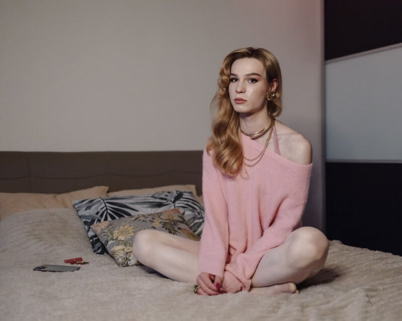A person with long, wavy blonde hair wearing a loose pink sweater sits cross-legged on a bed with patterned pillows, looking at the camera in a softly lit bedroom.