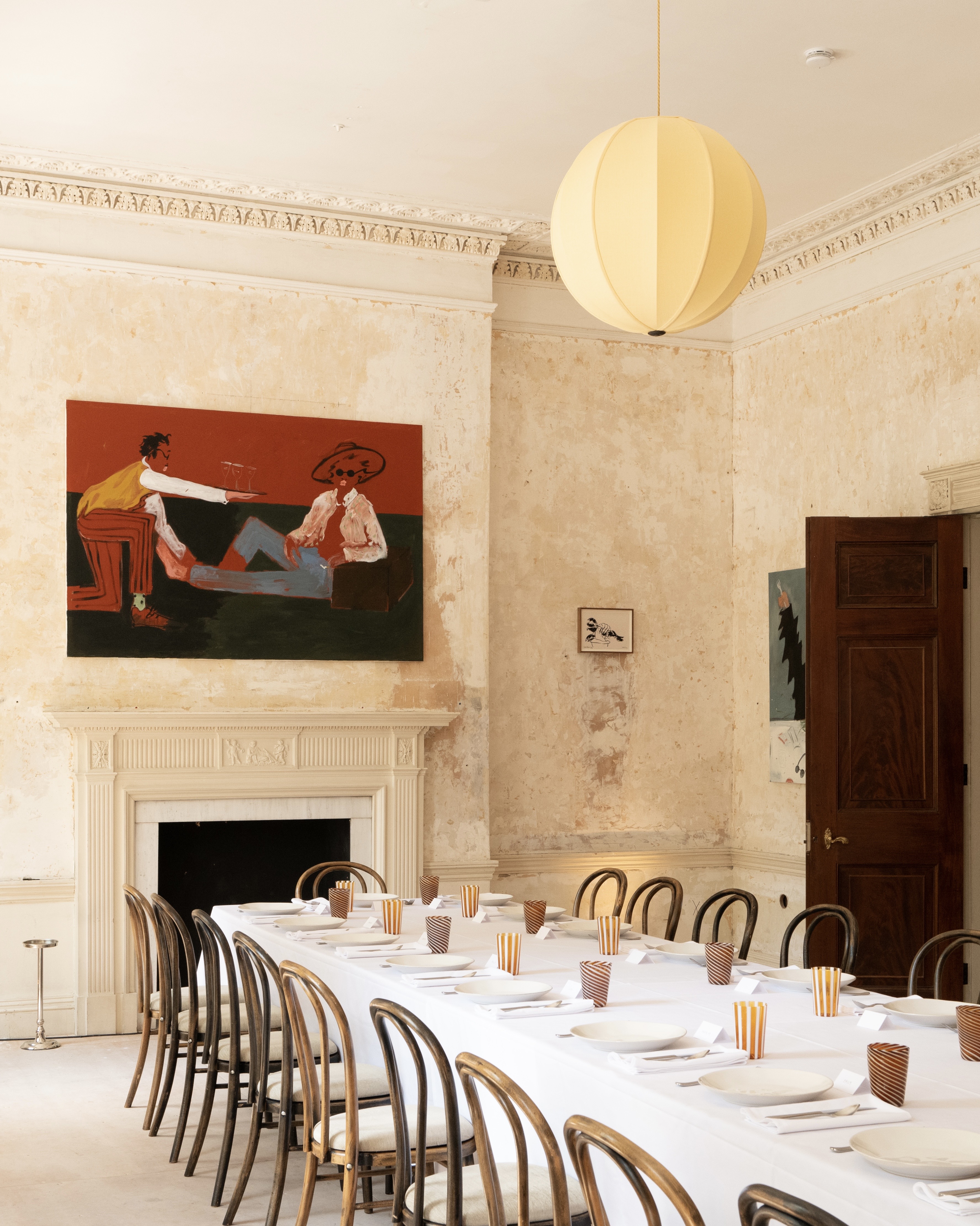 A long dining table set with white plates and striped glasses, surrounded by bentwood chairs, in a room with a large painting above a fireplace and textured walls.