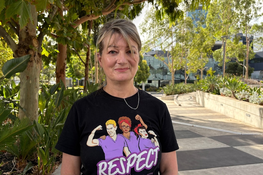 A woman has blonde hair tied up, she wears a black tshirt with three caricatured nurses