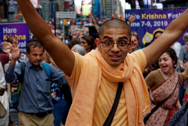 A man in orange robes with uplifted arms smiles amid a lively crowd in an urban setting, with "Hare Krishna Festival" banners visible in the background.