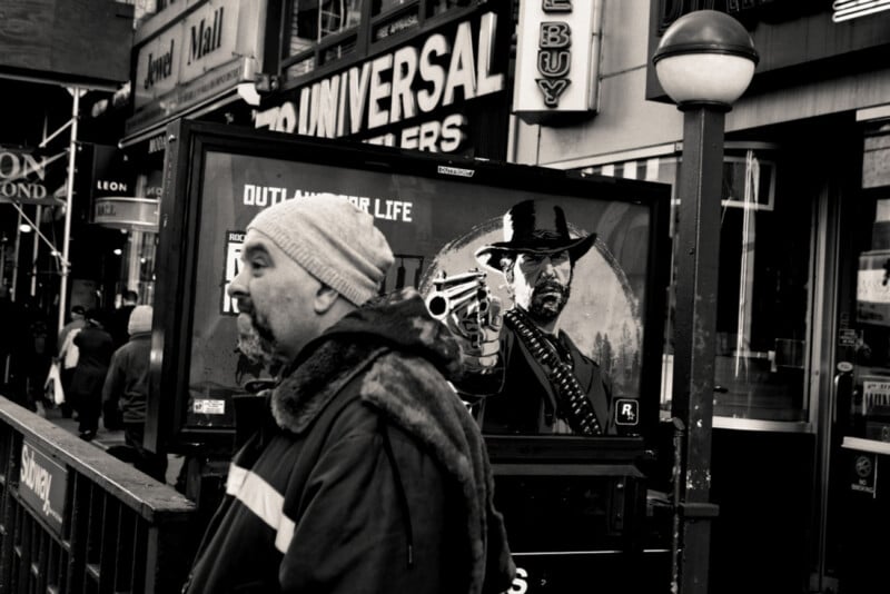 A man in a winter coat and hat walks past a subway entrance, with a large poster behind him showing a cowboy aiming a gun. The scene is busy with storefronts and people in the background.