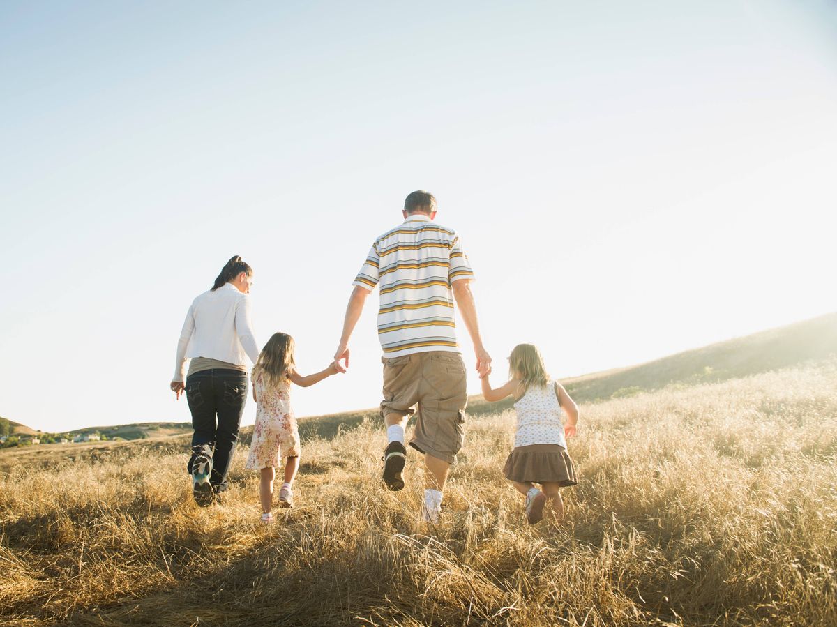 A family consisting of two adults and two children are walking hand-in-hand across a grassy field, bathed in the warm light of the sun.