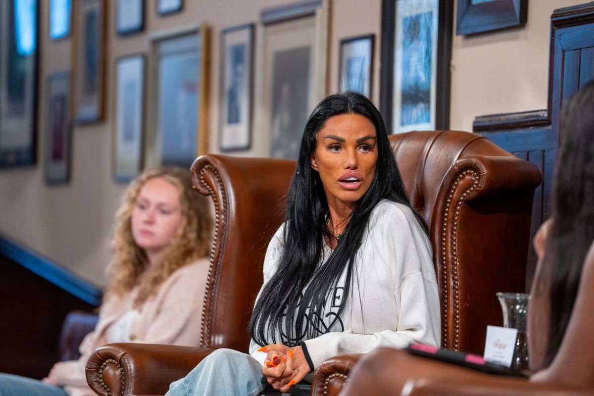 Two individuals, one seated with a glass and the other with a notebook, are engaged in conversation in a room adorned with framed pictures on the wall.