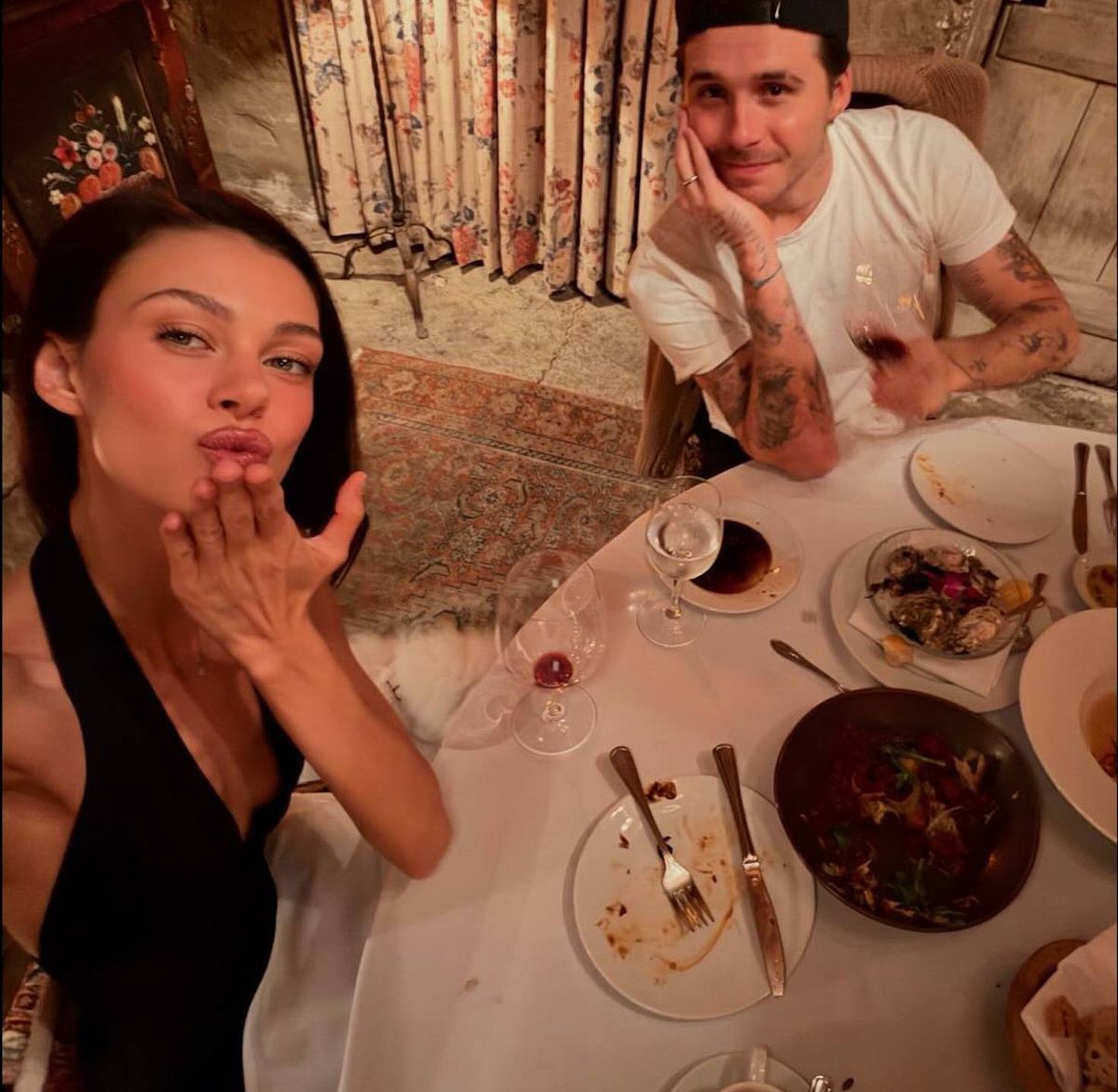 An indoor setting features two individuals seated at a table. The individual on the left is gazing upward with their hand resting on their chin, while the individual on the right is leaning on the table, their hand on their chin, and looking directly at the camera. The table is set with plates, utensils, and various dishes.