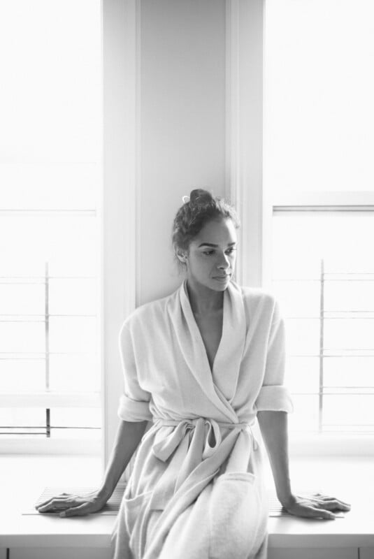 A woman in a bathrobe sits on a windowsill, gazing thoughtfully to the side. The scene is in black and white, with soft light streaming through the windows behind her.
