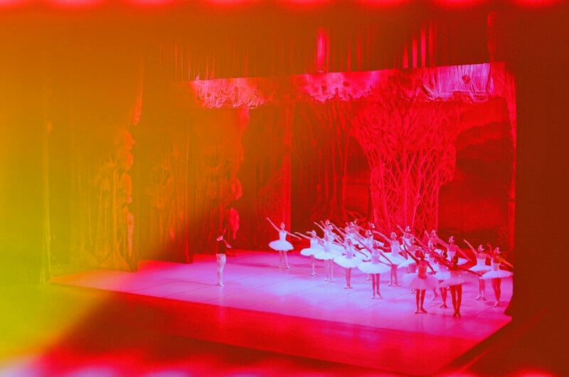 A group of ballet dancers perform on a brightly lit stage with elaborate red and pink forest-themed scenery, bathed in dramatic red and yellow lighting.