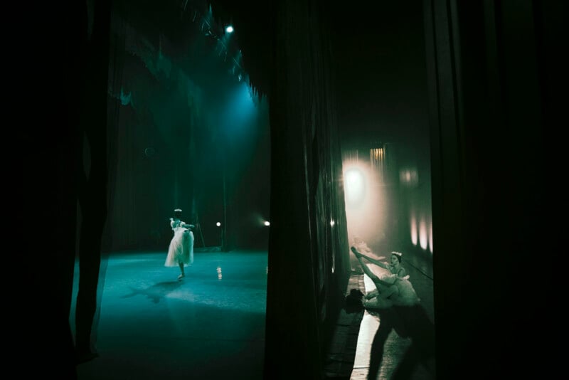 A ballerina dances under stage lights on an empty stage while another dancer sits stretching in the dimly lit wings, partially hidden behind dark curtains. The scene has a dramatic, atmospheric lighting.