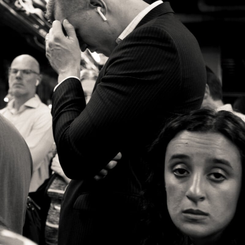 A crowded subway scene in black and white shows a man in a suit holding his head in stress, while a woman in the foreground looks tired and disheartened. Other commuters stand around them with serious expressions.