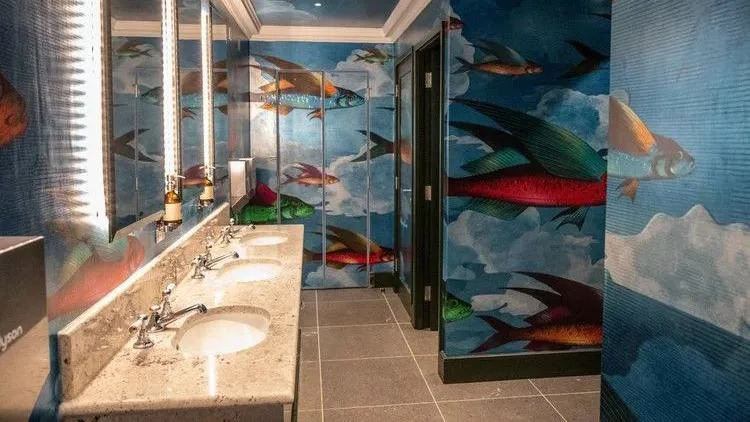 An underwater scene with fish swimming in a blue ocean all over the walls of the men's toilets. There is a marble wash basin in the well-lit room.