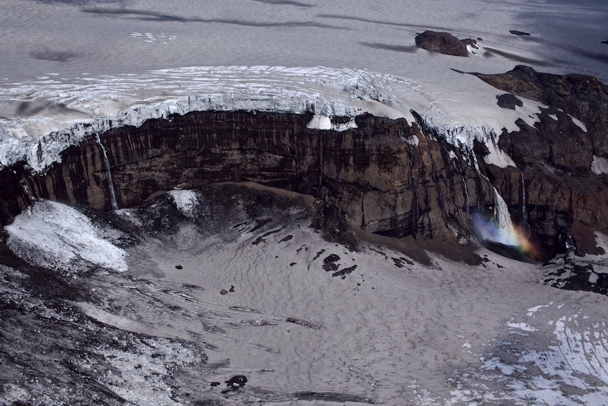 A photograph of a snowy plateau that forms a waterfall at a cliff's edge, a rainbow forming in the mist at the bottom.
