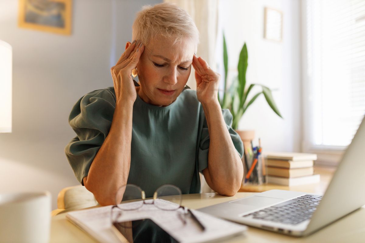 Senior female teacher sitting at desk massaging her temples suffering terrible headache after conducting online lesson from her cabinet. Mature businesswoman tired after hard work day