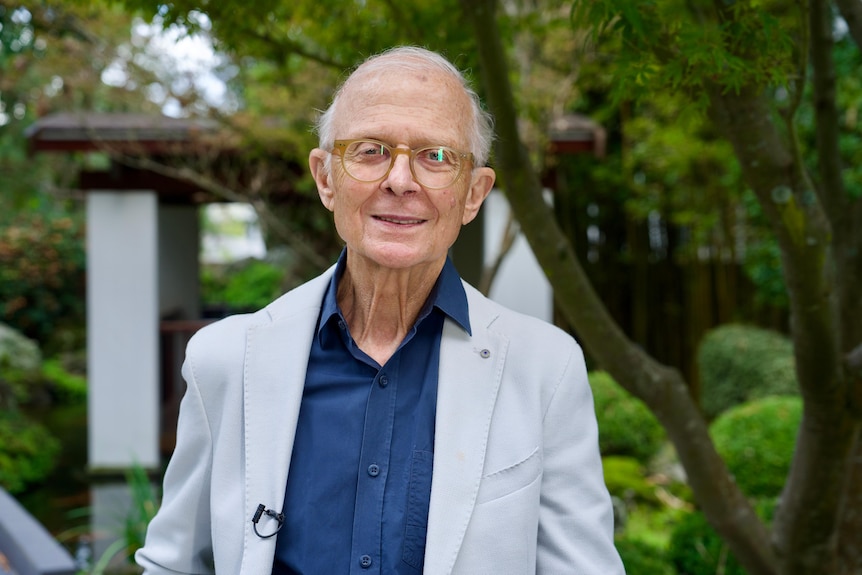 An older man with light white hair and glasses stands in a light grey suit and blue shirt with trees in the background.