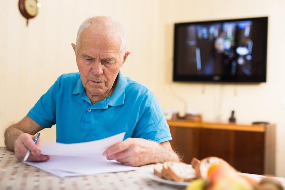 A senior man with white hair and a blue polo shirt sits at a table, holding a pen in his right hand and reviewing white papers. A stack of papers is on the patterned table in front of him. To his right, there's a plate with slices of bread and some fruit. In the blurry background, a wall-mounted television and a wooden cabinet are visible.