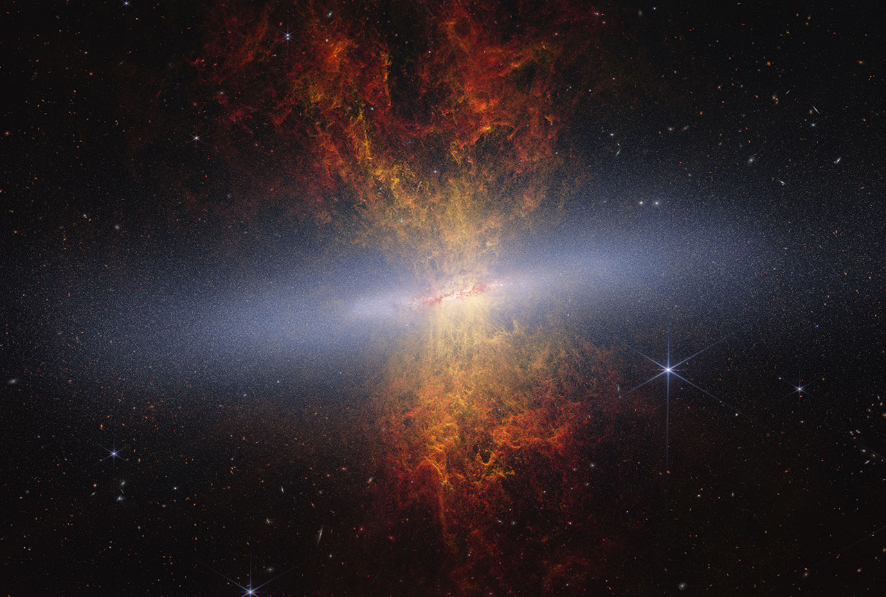 Composite image of edge-on spiral starburst galaxy Messier 82 as observed by NASA's James Webb and Hubble space telescopes. Hourglass-shaped plumes of gas are shooting outward from above and below a bright blue-white, disk-shaped center. The plumes are yellow near the galaxy’s bright center, denoting areas of ionized hydrogen gas as observed by Hubble, and gradually become redder as you move farther away. Messier 82 is set against the black background of space, which has many distant galaxies that appear as small white and orange spirals, ovals, and points of light. Toward the right of Messier 82 is a blue-white star with eight-pointed diffraction spikes that are characteristic of Webb.