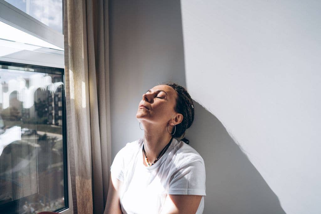 Woman in white t-shirt and hoop earrings, eyes closed, leaning against a window.
