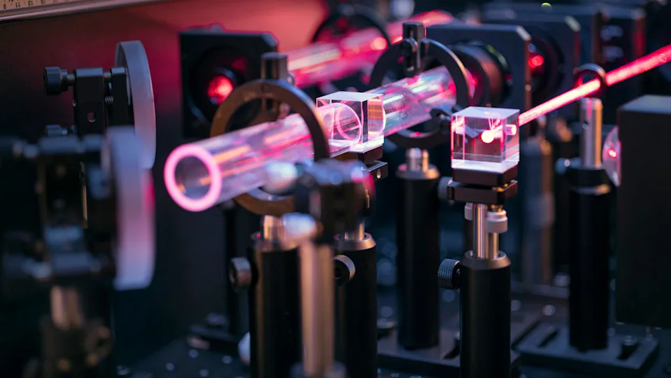 A close up of an optical table with various red lasers beamed across lenses and mirrors