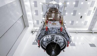 A large spacecraft suspended vertically inside a clean room, viewed from below. The spacecraft’s cylindrical base, wiring, and reflective insulation are visible, along with solar array panels folded along its sides. Bright overhead lights and white walls surround the vehicle. Photo credit: NASA/Sydney Rohde (Rocz)