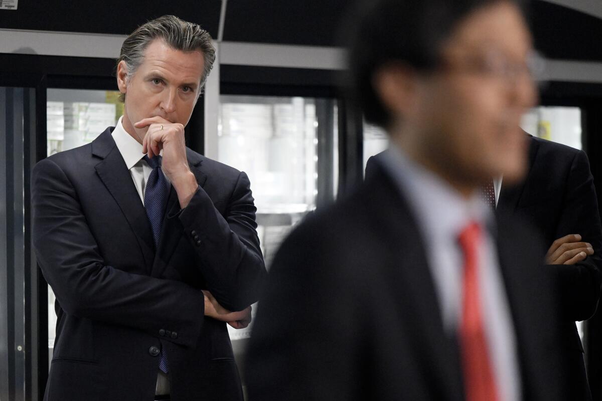 Gov. Gavin Newsom stands with one hand on his chin, watching someone else speak.